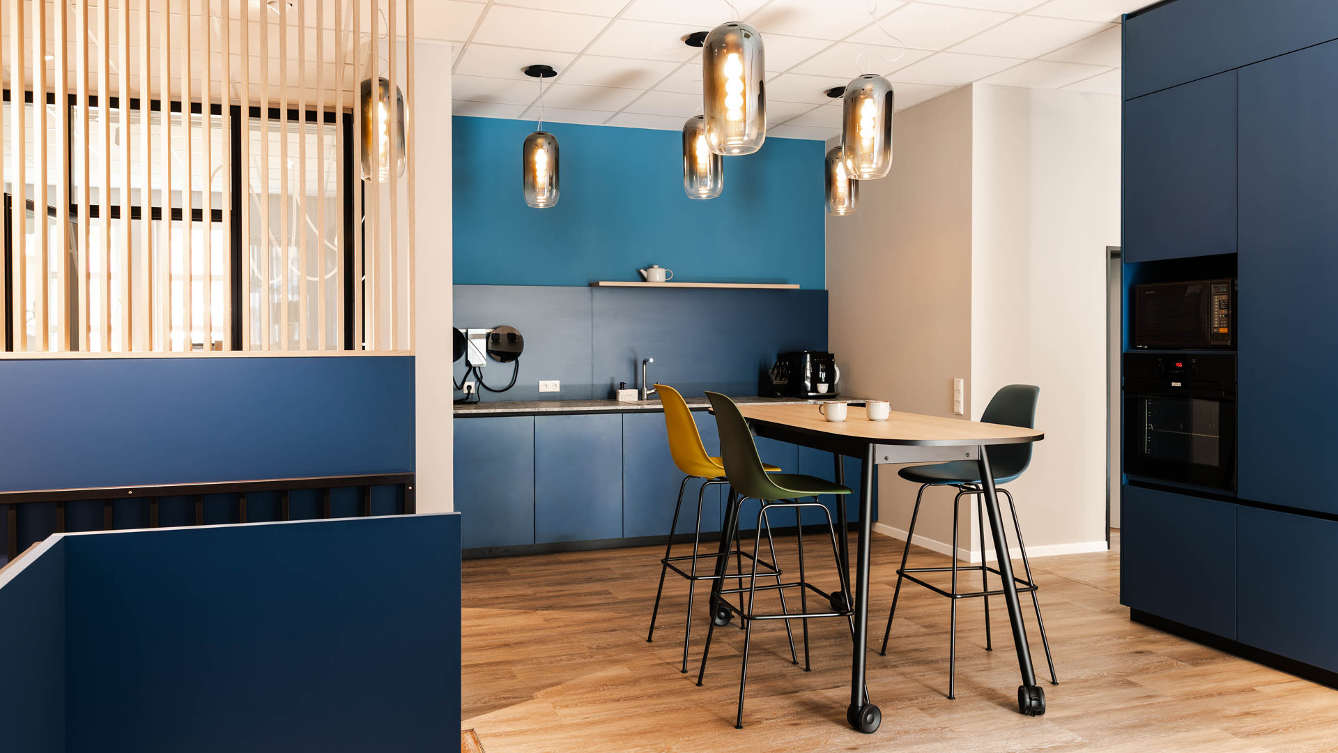 Kaffeebar und Aufenthaltsbereich von Apondium Apotheken in Düsseldorf mit dunkelblauen Einbauten, Holzlamellen und flexiblem Hochtisch