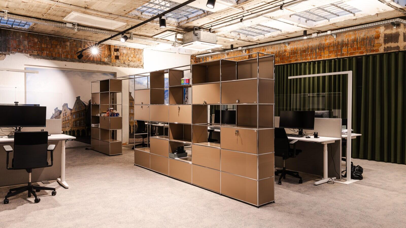 Offener Arbeitsbereich der Volksbank im Münsterland mit modularen Regalen, höhenverstellbaren Schreibtischen und sichtbarer Bestandsarchitektur.